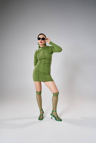 Pełnowymiarowe zdjęcie stylowej kobiety w zielonym stroju i dotykającej okularów jedną ręką - Zdjęcie, obraz