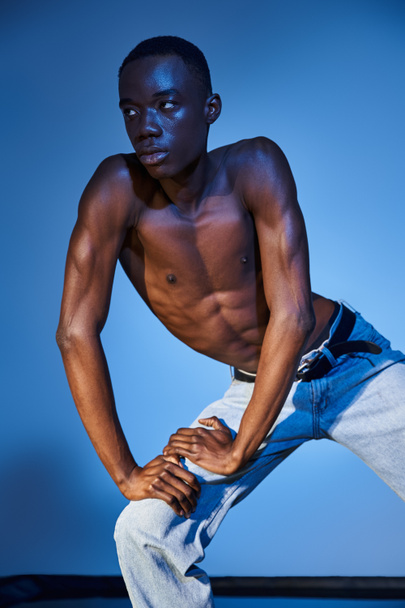 μοντέρνο αφροαμερικανός άνδρας σε κομψό τζιν ποζάρουν σε μπλε υδαρές φόντο και κοιτάζοντας μακριά - Φωτογραφία, εικόνα