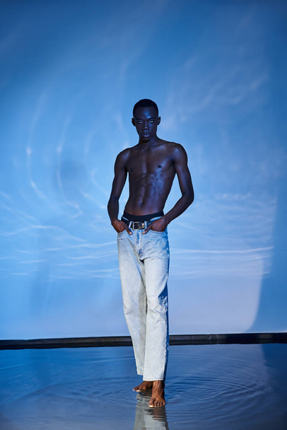 kuszący afrykański mężczyzna bez koszuli w modnych dżinsach patrzący w kamerę na wodnistym tle - Zdjęcie, obraz
