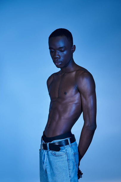 uomo afroamericano senza maglietta in jeans alla moda in posa su sfondo acquoso blu e guardando altrove - Foto, immagini