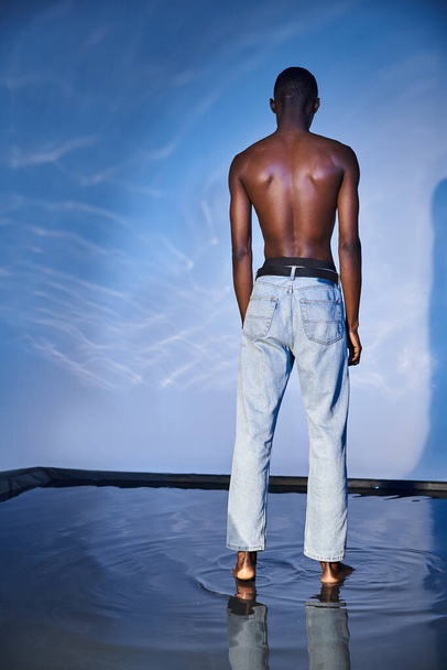vue arrière de l'homme afro-américain torse nu à la mode en jeans tendance sur fond bleu aqueux - Photo, image