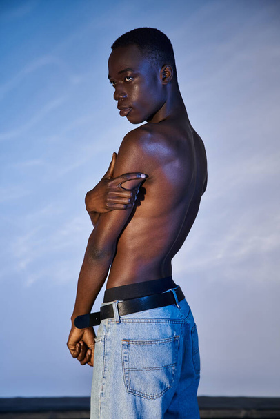 без сорочки афроамериканський чоловік в стильних джинсах позує на синьому водяному фоні і дивиться в сторону - Фото, зображення