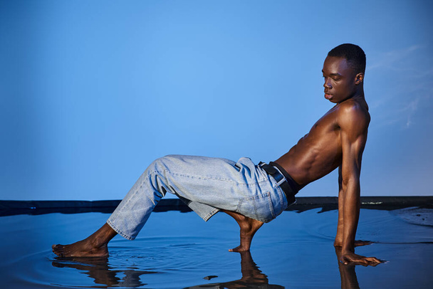 přitažlivý bez košile africký Američan muž v džínách pózuje v pohybu a dívá se jinam na modré pozadí - Fotografie, Obrázek