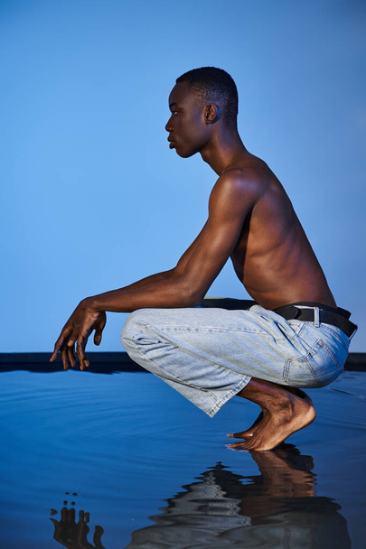 shirtloze stijlvolle Afrikaans-Amerikaanse man in trendy jeans hurkend en wegkijkend op waterige achtergrond - Foto, afbeelding