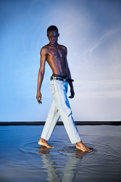 alluring young african american man in stylish jeans posing topless on watery blue background - Photo, Image