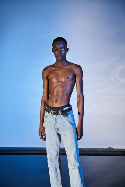 bez koszulki stylowy Afrykański Amerykanin w nowoczesnych dżinsach i patrząc na aparat fotograficzny na niebieskim tle - Zdjęcie, obraz