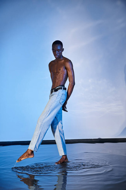 atraktivní mladý africký Američan muž ve stylové džíny pózovat nahoře bez na vodnaté modré pozadí - Fotografie, Obrázek