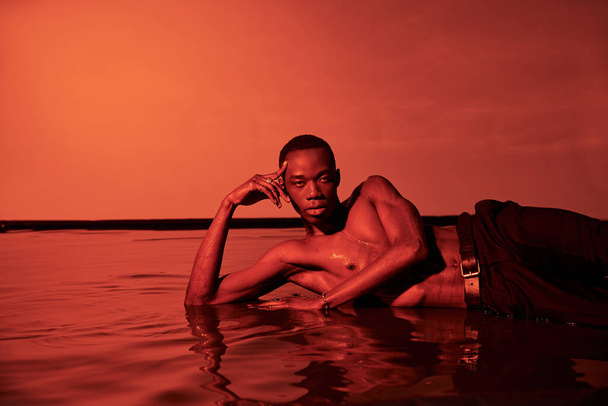 jovem atraente afro-americano homem posando topless e reclinado na superfície da água em luzes vermelhas - Foto, Imagem