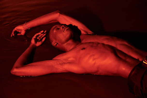 sexy séduisante sans chemise homme afro-américain couché sur la surface de l'eau avec les yeux fermés dans les lumières rouges - Photo, image