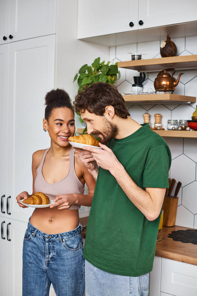 good looking joyous diverse couple in comfy homewear holding plates with croissants in kitchen - Photo, Image