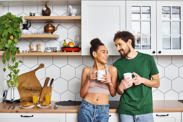 приваблива весела різноманітна пара в домашньому одязі, насолоджуючись гарячою кавою і дивлячись один на одного - Фото, зображення