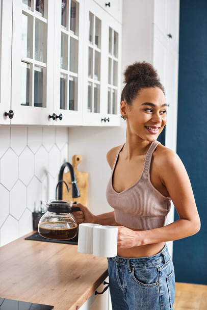 joyous african american beautiful woman in cozy homewear holding coffee and looking away in kitchen - Photo, Image