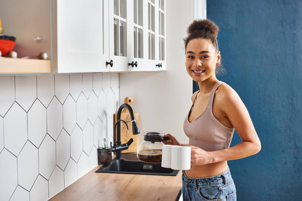 fröhlich ansprechende afrikanisch-amerikanische Frau in Homewear hält Kaffee und lächelt in die Kamera - Foto, Bild