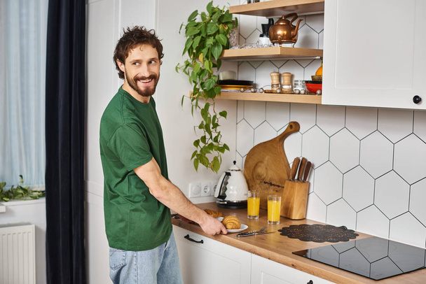 красивий веселий чоловік в повсякденному одязі готується їсти круасани і дивитися на кухню - Фото, зображення