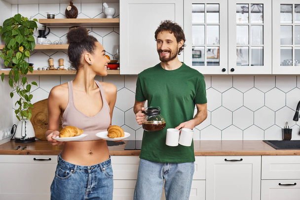 veselý multiraciální pár v domácím oblečení připravuje snídani s kávou a croissanty - Fotografie, Obrázek