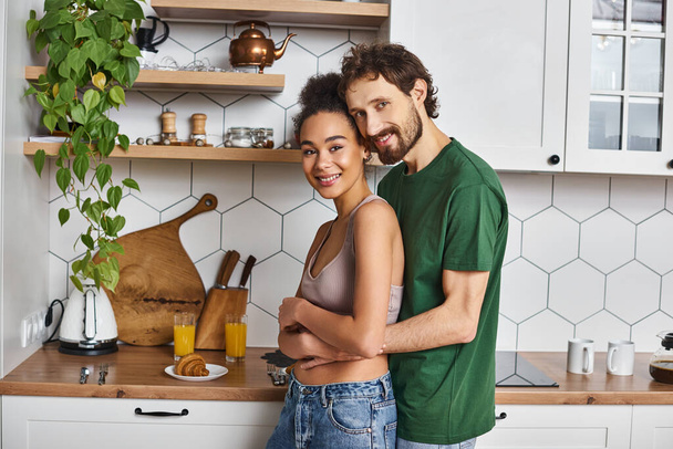 veselý dobře vypadající multikulturní pár v homewear objímání láskyplně a úsměv na kameru - Fotografie, Obrázek