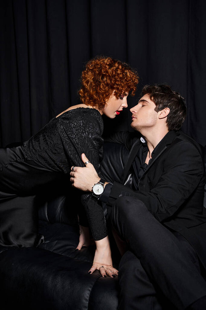 houkutteleva poikaystävä ja tyttöystävä tyylikkäissä vaatteissa halaamalla rakastavasti mustalla sohvalla, seksikäs pari - Valokuva, kuva