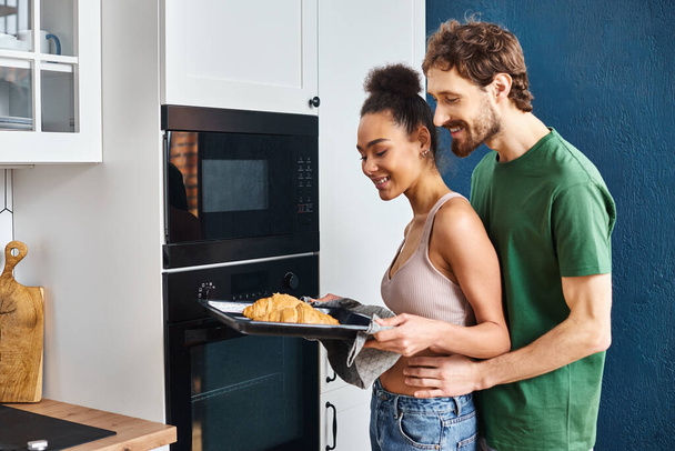 alegre pareja multicultural en ropa de casa utilizando el horno para hornear deliciosos croissants para el desayuno - Foto, imagen