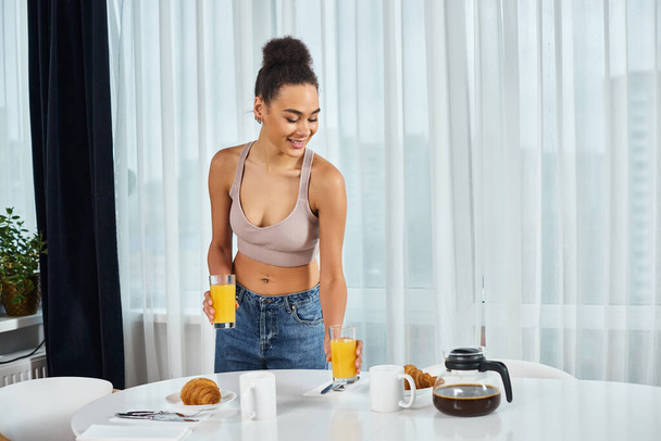 自宅で朝食のためにオレンジジュースを提供するホームウェアで若いアフリカ系アメリカ人女性を魅了 - 写真・画像