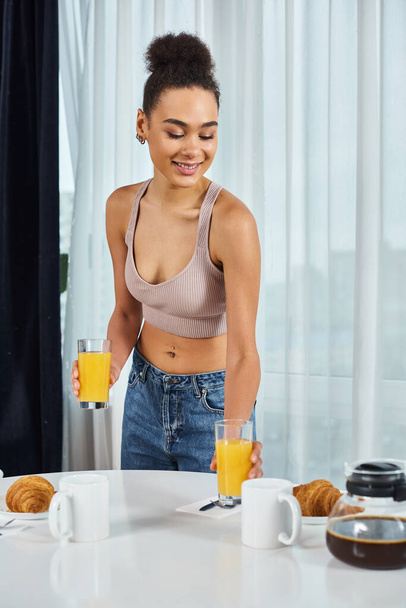 atraktivní veselá africká americká žena v útulném oblečení sloužící čerstvé pomerančové šťávy, zatímco doma - Fotografie, Obrázek