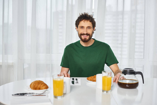 Χαρούμενος ελκυστικός άντρας με περιστασιακή ενδυμασία κάθεται στο τραπέζι κατά τη διάρκεια του πρωινού και χαμογελά στην κάμερα - Φωτογραφία, εικόνα