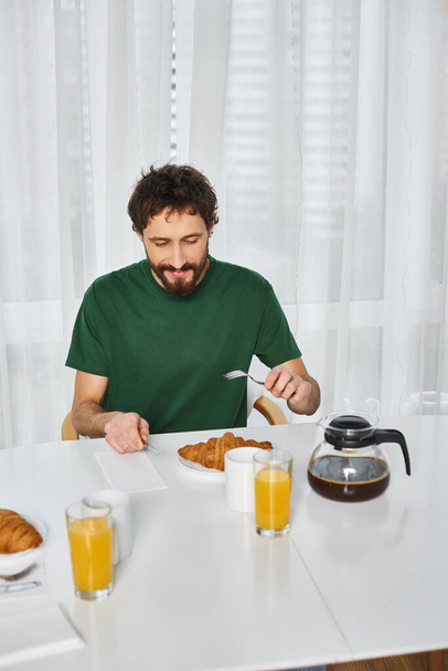 Güzel görünümlü, rahat ev elbiseli, keyifli bir adam lezzetli bir kahvaltı yapıyor ve evde mutlu bir şekilde gülümsüyor. - Fotoğraf, Görsel