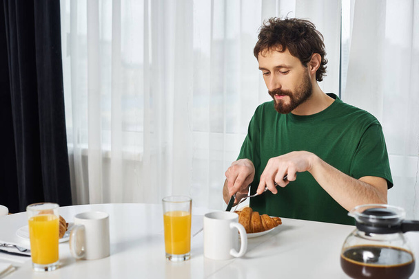 pohledný muž v ležérní zelené tričko řezání lahodné croissant s nožem při snídani doma - Fotografie, Obrázek