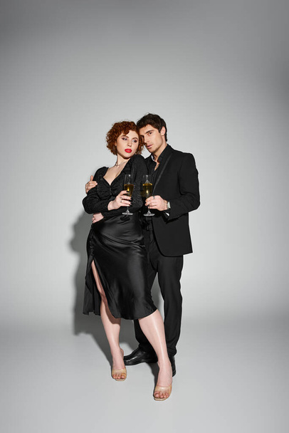 mooie vrouw in zwart jurk knuffelen haar knappe vriendje op grijze achtergrond, champagne in handen - Foto, afbeelding