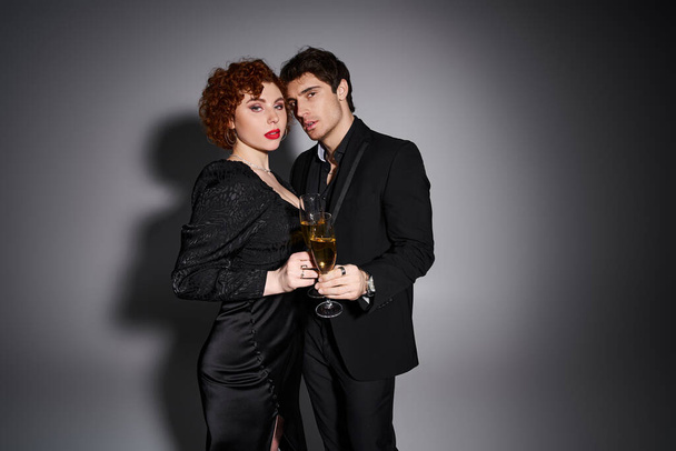 сексуальная красивая пара в черном платье обнимающая и смотрящая в камеру с шампанским в руках - Фото, изображение