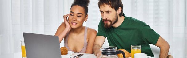 atractiva pareja multicultural alegre en ropa de casa viendo películas durante el desayuno, pancarta - Foto, Imagen