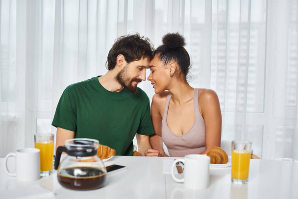joyous good looking multicultural couple posing lovingly together during their breakfast at home - Photo, Image