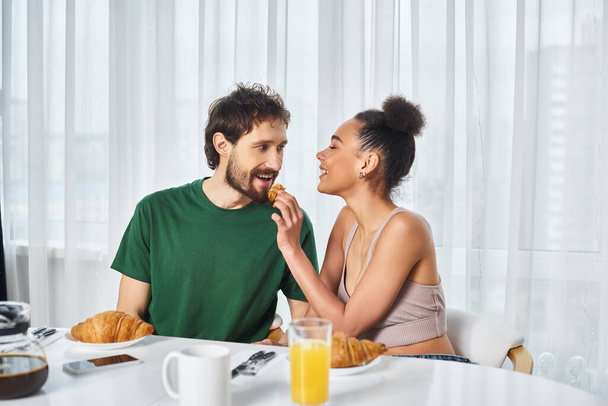 İç çamaşırlı neşeli Afro-Amerikan kadın yakışıklı sevgilisine kruvasan yediriyor. - Fotoğraf, Görsel