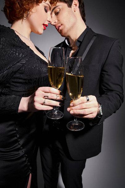 verleidelijke vrouw in zwart jurk knuffelen haar knappe vriendje op grijze achtergrond, champagne in handen - Foto, afbeelding
