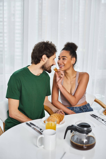 cheerful beautiful african american woman feeding delicious croissant to her loving boyfriend - Photo, Image