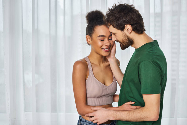 atractiva pareja interracial alegre en acogedora ropa de casa abrazándose amorosamente y sonriendo felizmente en casa - Foto, Imagen