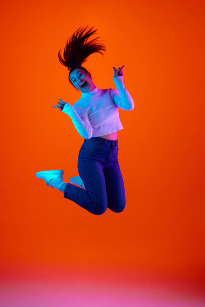 Heyecanlı genç kadının tam boy portresi neon ışıkta pembe-turuncu arkaplana karşı mutluluk ve neşe içinde zıplıyor. Kendini ifade etme kavramı, güzellik ve moda, enerji. Ad - Fotoğraf, Görsel