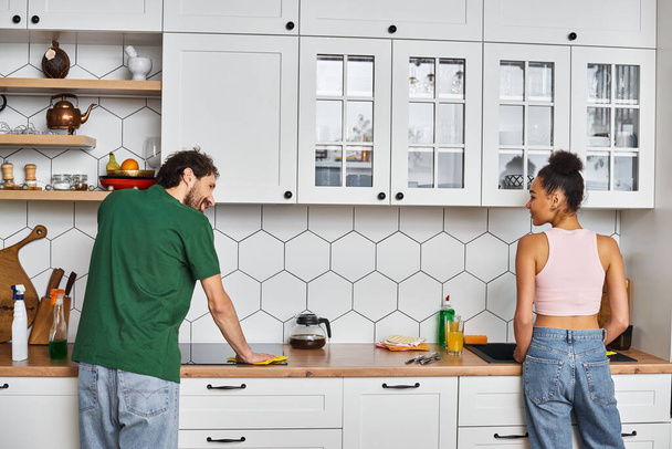 ελκυστικό χαρούμενο πολυπολιτισμικό ζευγάρι σε ζεστή ενδυμασία καθαρίζει κουζίνα μετά το πρωινό μαζί - Φωτογραφία, εικόνα