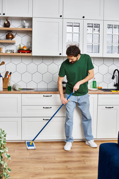красивый веселый мужчина в удобной повседневной одежде, используя швабру для мытья пола, находясь дома на кухне - Фото, изображение