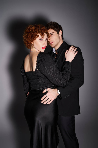pareja apasionada de buen aspecto en trajes chic negro abrazando y mirando a la cámara en el fondo gris - Foto, imagen