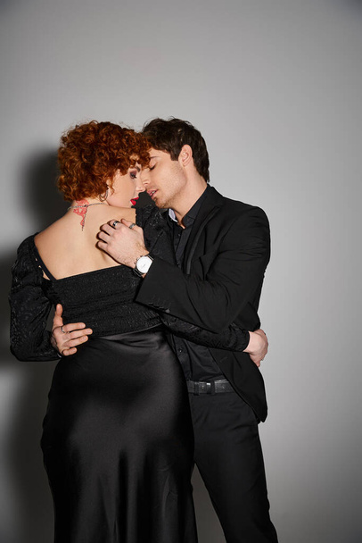 соблазнительная сексуальная пара в черных элегантных одеждах целуется и обнимается с любовью на сером фоне - Фото, изображение