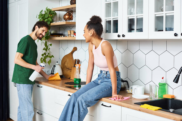cheerful african american woman sitting on counter and looking at her boyfriend cleaning kitchen - Photo, Image