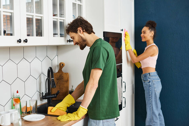 Весёлая мультикультурная красивая пара в домашней одежде делает свою работу во время весенней уборки. - Фото, изображение