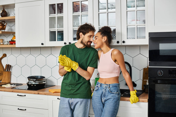 atractiva pareja interracial alegre en ropa de casa disfrutando el uno del otro durante la limpieza de primavera - Foto, Imagen