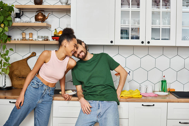appealing joyous multiracial couple in homewear spending time together while in kitchen at home - Photo, Image