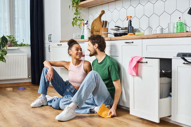 appealing multiracial joyous couple in homewear sitting on floor and smiling at each other - Photo, Image