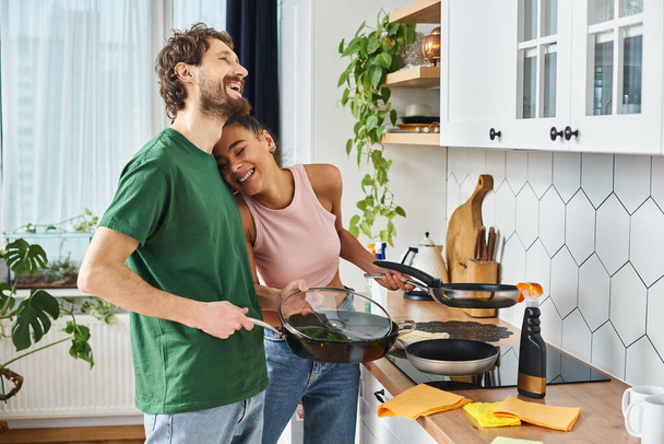 весела приваблива мультикультурна пара в домашньому одязі тримає сковорідки в руках і весело посміхається - Фото, зображення