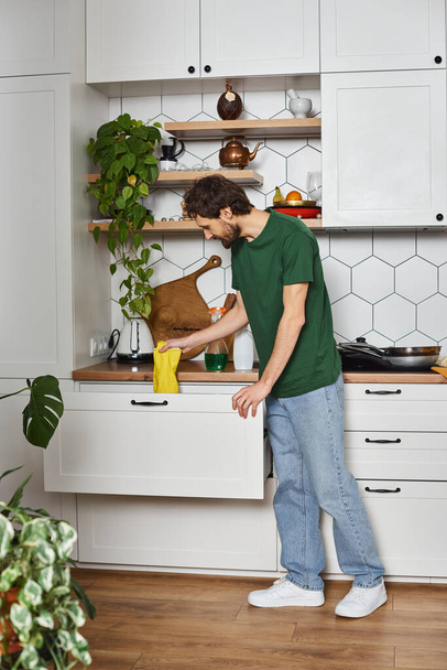 Красивый мужчина в темно-зеленой уютной футболке делает домашнюю работу и держит тряпку в руке во время весенней уборки - Фото, изображение