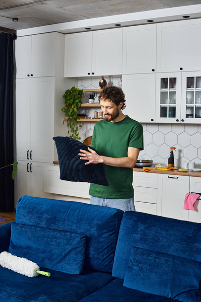 attractive joyous man in casual green t shirt holding pillow in his hands while cleaning at home - Photo, Image