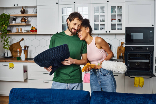 приваблива весела мультикультурна пара в зручному домашньому одязі чистить пил вдома і щасливо посміхається - Фото, зображення