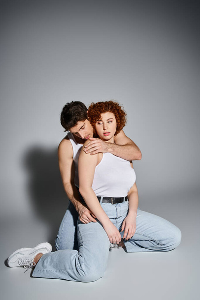 alluring man and woman in blue jeans sitting on floor together and hugging warmly, sexy couple - Photo, Image